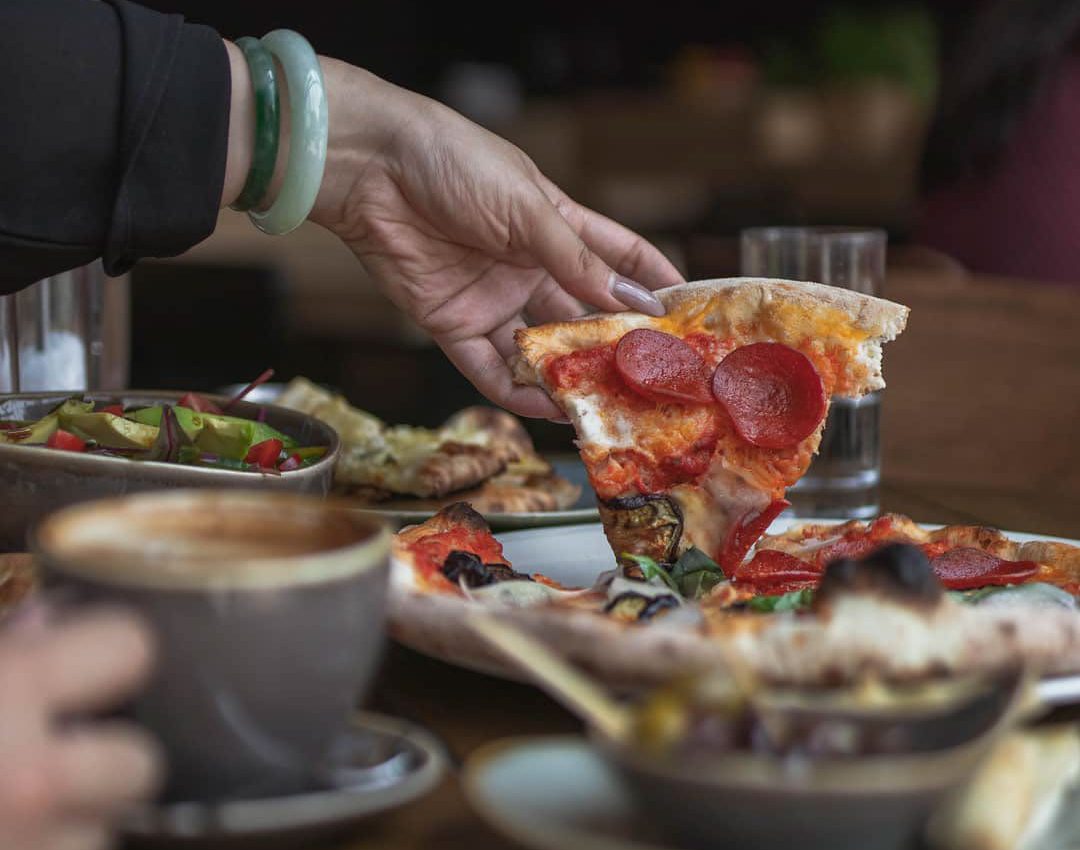 Woman grabbing pizza with pepperoni, peppers and mushroom at Zzetta. She has coffee, a salad, and dessert on the side.